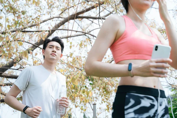 Long hair woman wearing old rose color sport bra running in the park in autumn, a man in white t-shirt running behind her.