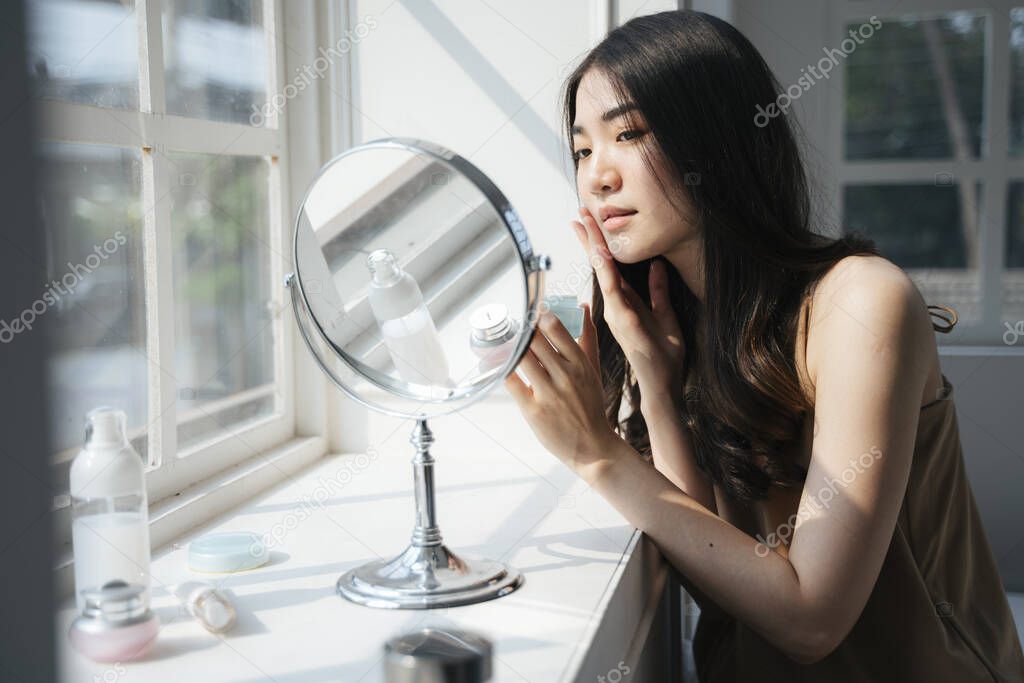 Wavy long hair girl apply the facial cream on her face gently while sitting in front of the mirror next to the window of her bedroom.