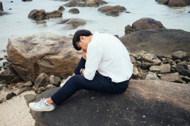 Beyaz tişörtlü siyah adam denizde kayanın üzerinde oturuyor ve başını dizine dayıyor..