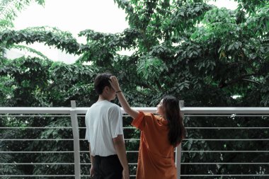 Turuncu tişörtlü kız bir adamın saçına dokunuyor. Ağacın korkuluğunda dikiliyorlar..