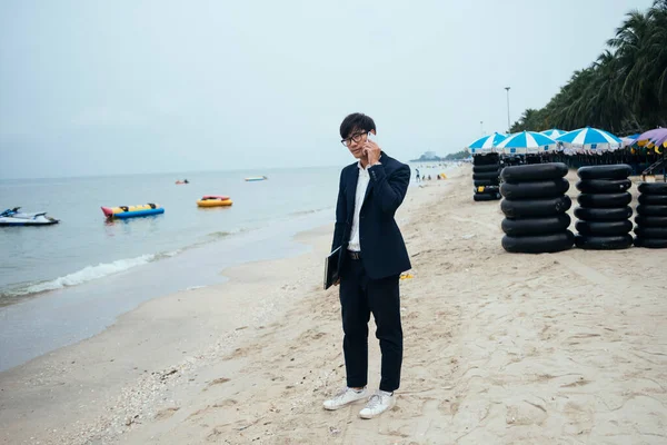 Black hair guy in dark suit standing on the beach and talking with his customer on the phone under unclear sky.