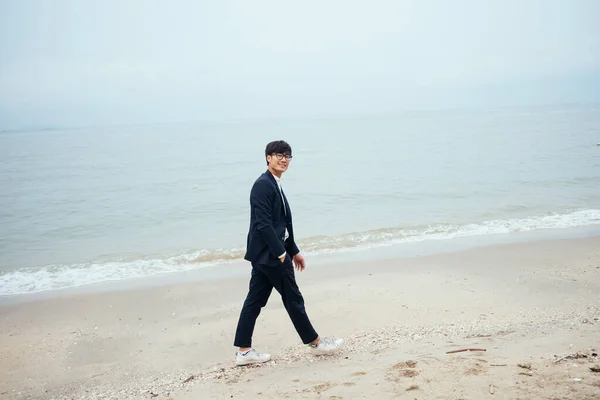 Black hair guy in dark suit walking along the beach with unclear sky look at the camera.