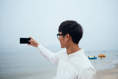 Beyaz tişörtlü siyah adam kasvetli gökyüzü ile sahilde duruyor ve akıllı telefonla denizin fotoğrafını çekiyor..