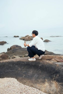 Beyaz gömlekli siyah adam denizde kayanın üzerinde elinde şişeyle oturuyor, denize bak..