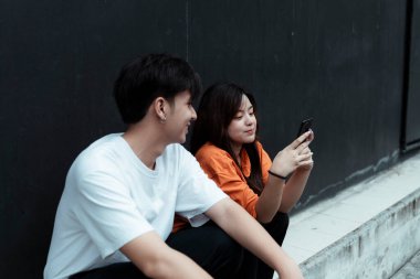 Genç bir çift siyah duvarda oturuyor, bir kız telefonunu erkek arkadaşına gösteriyor ve gülümsüyor..