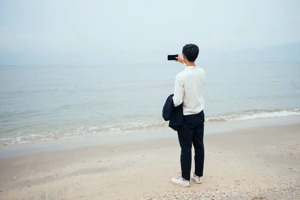 Black hair guy in white shirt standing on the beach with unclear sky and take a photo of the sea with smartphone, full shot.