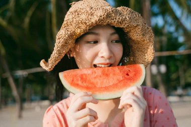 Kahverengi şapkalı bir kız sahilde dururken karpuz yiyor..