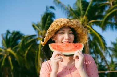 Kahverengi şapkalı bir kız sahilde dururken karpuz yiyor..