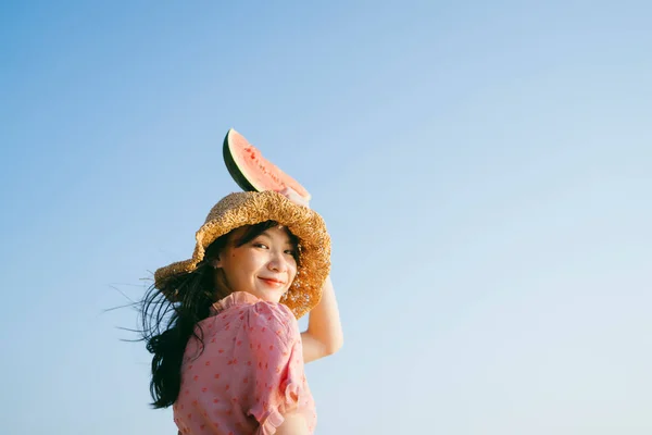 Cute girl in brown hat raise the watermelon at her head and smile under blue sky.