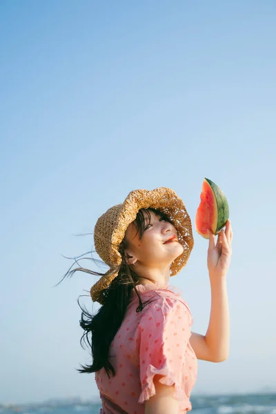 Kahverengi şapkalı tatlı kız kafasında karpuzu kaldırıyor ve mavi gökyüzünün altında gülümsüyor..