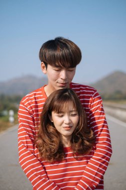 Bir adam, dağ manzaralı yolda dikilirken kız arkadaşına arkadan sarılıyor..