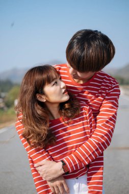 Bir adam kız arkadaşını sırtından kucaklıyor ve dağ manzaralı yolda dikilirken birbirlerine bakıyor..