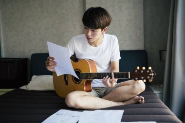 Genç Taylandlı gitarist akustik gitar çalışıyor ve yatak odasında şarkı sözü kağıdı notaları okuyor..
