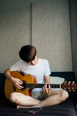 Genç Taylandlı gitarist akustik gitar çalarken akıllı telefondan kulaklıklı şarkılar dinliyor..