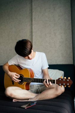 Genç Taylandlı gitarist akustik gitar çalarken akıllı telefondan kulaklıklı şarkılar dinliyor..