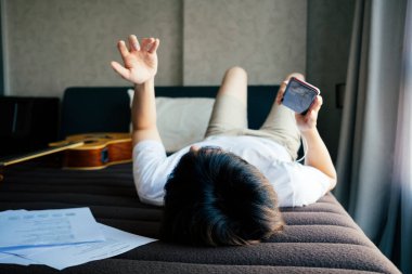 Genç Taylandlı gitarist yatağa uzanmış akıllı telefondan şarkılar dinliyor ve hava gitarıyla pratik yapıyor..