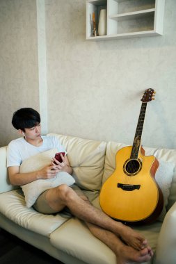 Genç Taylandlı gitarist kanepeye uzanıp akıllı telefon ve kulaklıkla müzik dinliyor..