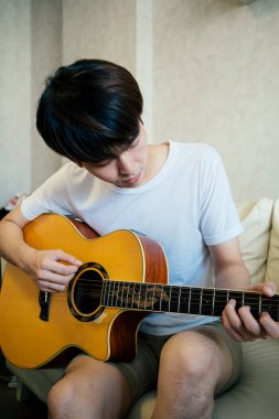 Beyaz tişörtlü genç Taylandlı gitaristin oturma odasındaki bir kanepede akustik gitar çalmasını kapat..