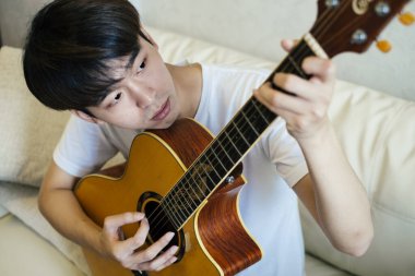 Genç Taylandlı gitarist evdeki koltukta akustik gitar çalıyor..
