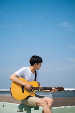 Genç Taylandlı gitarist bir metroda oturmuş açık havada çatıda akustik gitar çalıyor..