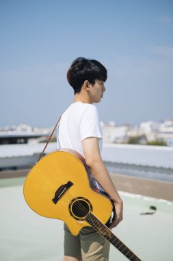 Sideview - Genç Taylandlı gitarist çatıda akustik gitar taşıyor.