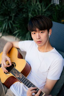 Genç Taylandlı gitaristin portresi kulaklıkla müzik dinlemek ve akustik gitar çalmak..