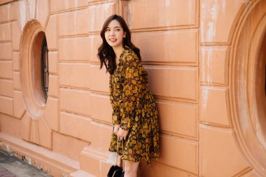 Sarı elbiseli tatlı kıvırcık saçlı kız turuncu duvara yaslanıyor ve siyah arkasını yatay olarak tutuyor..