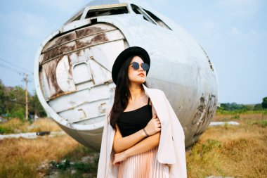 Pembe takım elbiseli, siyah şapkalı, güneş gözlüklü siyah uzun saçlı kız terk edilmiş bölgedeki harabe uçağın önünde duruyor..