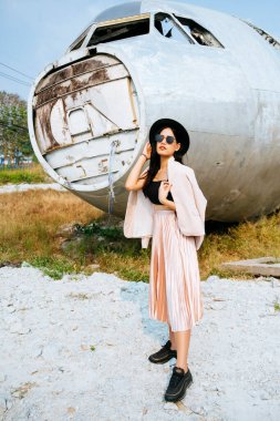 Siyah uzun saçlı kız, siyah şapkalı, pembe etekli ve enkaz uçağının önünde dikilirken takım elbisesini omuzlarına giymiş..