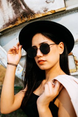 Siyah spagetti kayışlı, güneş gözlüğü ve şapka takan siyah uzun saçlı kızın orta boy portresi. Şapkasını al ve şık bir kız gibi elbisesini omzuna as..