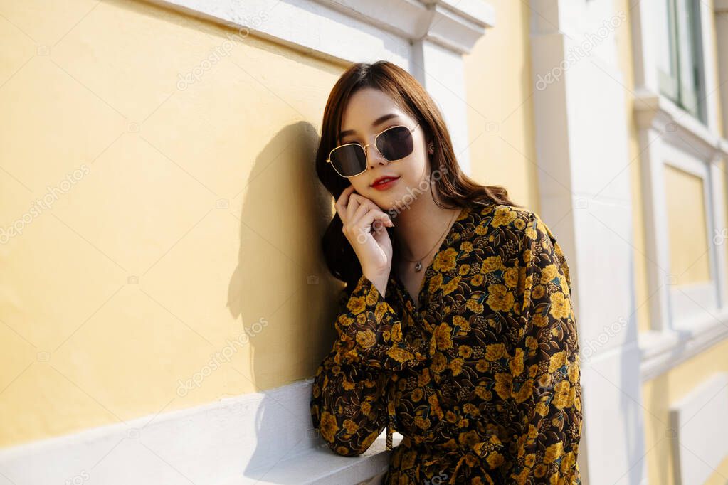 Cute curly long hair girl in yellow dress and sunglasses standing at the yellow and white building, look at the camera, horizontal.