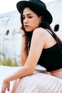 Siyah ve pembe elbiseli uzun saçlı kadın siyah şapkalı ve enkaz uçağının yanında oturuyor..