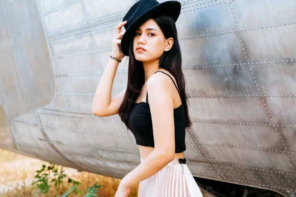Long hair woman in black and pink clothes wearing black hatstanding and sitting next to the wreck plane.