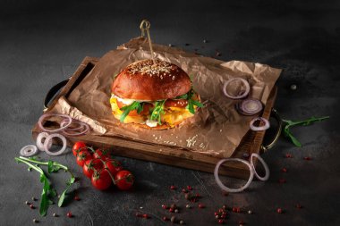 big american chicken curry burger on a black background. cutlet with cheese, tomato, onion and arugula on a wooden tray, side view