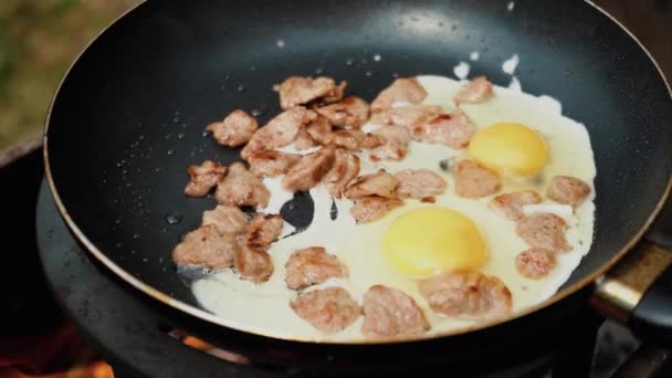 Primer plano de huevo roto cae en la sartén. Huevos y tocino, hierba verde sobre el fondo borroso, cocinado en la parrilla, desayuno de verano, comer en la naturaleza - Concepto Naturaleza y Ecología. — Vídeo de stock