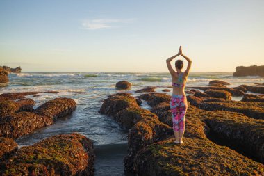 Gelgit sırasında büyük bir taşın üzerinde duran kadın. Genç bir kadın plajda namaste Mudra ile kollarını kaldırıyor. Gün batımı zamanı. Okyanus manzaralı. Bali 'de yoga inzivası, Mengening plajı, Endonezya. Arkadan görüntüle.