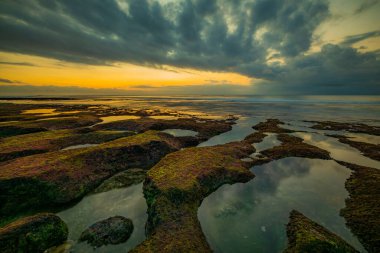 İnanılmaz bir deniz manzarası. Taşları ve taşları olan bir sahil. Dalgalar çekildi. Temiz su. Suda gökyüzü yansıması. Hava bulutlu. Kapama hızı yavaş. Yumuşak odaklanma. Ufukta güneş ışığı görünüyor. Balangan Sahili, Bali, Endonezya
