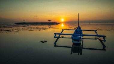 Deniz Burnu. Balıkçı teknesi Jung. Gün doğumu manzarası. Okyanustaki yapay bir adada çardaklar. Su yansıması. Güneş ufukta görünüyor. Kapama hızı yavaş. Yumuşak odaklanma. Sanur Plajı, Bali, Endonezya.