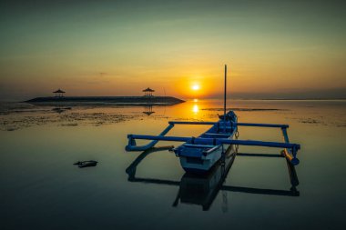 Deniz Burnu. Balıkçı teknesi Jung. Gün doğumu manzarası. Okyanustaki yapay bir adada çardaklar. Su yansıması. Güneş ufukta görünüyor. Kapama hızı yavaş. Yumuşak odaklanma. Sanur Plajı, Bali, Endonezya.