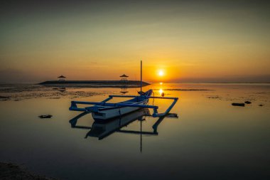 Deniz Burnu. Balıkçı teknesi Jung. Gün doğumu manzarası. Okyanustaki yapay bir adada çardaklar. Su yansıması. Güneş ufukta görünüyor. Kapama hızı yavaş. Yumuşak odaklanma. Sanur Plajı, Bali, Endonezya.