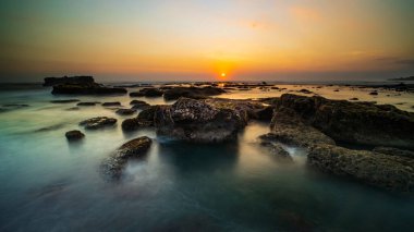 Arka plan için inanılmaz bir deniz manzarası. Taşları ve taşları olan bir sahil. Dalgalar çekildi. Hareket suyu. Gökyüzü açık. Güneş ufukta görünüyor. Peyzaj arka planı. Kapama hızı yavaş. Yumuşak odaklanma. Mengening Plajı, Bali, Endonezya