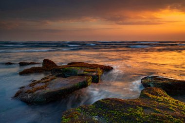Güzel deniz manzarası. Deniz yosunlarıyla kaplı taşlarla dolu bir sahil. Dalgalar çekildi. Doğanın bileşimi. Hareket suyu. Güneşli bulutlu bir gökyüzü. Kapama hızı yavaş. Yumuşak odaklanma. Mengening Plajı, Bali, Endonezya
