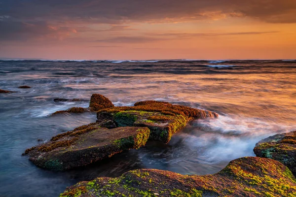 Güzel deniz manzarası. Deniz yosunlarıyla kaplı taşlarla dolu bir sahil. Dalgalar çekildi. Doğanın bileşimi. Hareket suyu. Güneşli bulutlu bir gökyüzü. Kapama hızı yavaş. Yumuşak odaklanma. Mengening Plajı, Bali, Endonezya