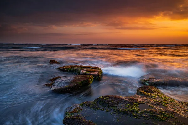 Güzel deniz manzarası. Deniz yosunlarıyla kaplı taşlarla dolu bir sahil. Dalgalar çekildi. Doğanın bileşimi. Hareket suyu. Güneşli bulutlu bir gökyüzü. Kapama hızı yavaş. Yumuşak odaklanma. Mengening Plajı, Bali, Endonezya