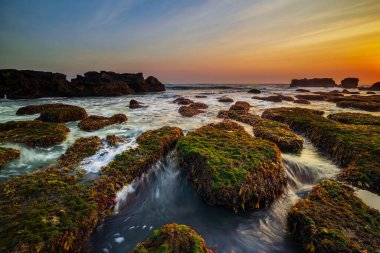İnanılmaz bir deniz manzarası. Hareket eden dalgalı okyanus. Düşük lide. Yeşil yosun ve yosunlarla kaplı taşlar. Doğa geçmişi kavramı. Gün batımı manzarası. Uzun pozlama. Yumuşak odaklanma. Mengening Plajı, Bali, Endonezya