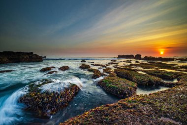 İnanılmaz bir deniz manzarası. Hareket eden dalgalı okyanus. Düşük lide. Yeşil yosun ve yosunlarla kaplı taşlar. Güneş ufukta görünüyor. Günbatımı manzara arka planı. Uzun pozlama. Yumuşak odaklanma. Mengening Plajı, Bali, Endonezya