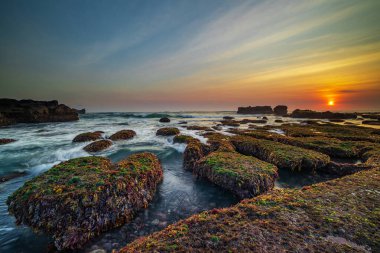 İnanılmaz bir deniz manzarası. Hareket eden dalgalı okyanus. Düşük lide. Yeşil yosun ve yosunlarla kaplı taşlar. Güneş ufukta görünüyor. Günbatımı manzara arka planı. Uzun pozlama. Yumuşak odaklanma. Mengening Plajı, Bali, Endonezya