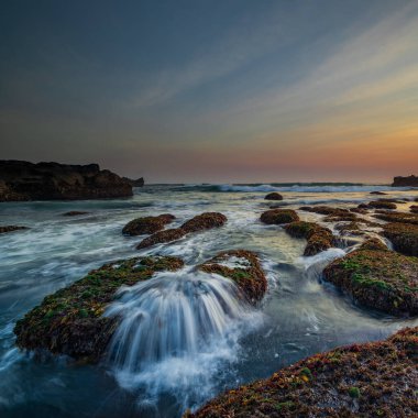 İnanılmaz bir deniz manzarası. Hareket eden dalgalı okyanus. Düşük lide. Yeşil yosun ve yosunlarla kaplı taşlar. Doğa geçmişi kavramı. Gün batımı manzarası. Uzun pozlama. Yumuşak odaklanma. Mengening Plajı, Bali, Endonezya