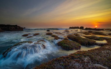İnanılmaz bir deniz manzarası. Hareket eden dalgalı okyanus. Düşük lide. Yeşil yosun ve yosunlarla kaplı taşlar. Güneş ufukta görünüyor. Günbatımı manzara arka planı. Uzun pozlama. Yumuşak odaklanma. Mengening Plajı, Bali, Endonezya