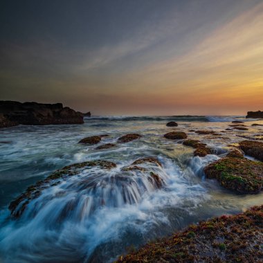 İnanılmaz bir deniz manzarası. Hareket eden dalgalı okyanus. Düşük lide. Yeşil yosun ve yosunlarla kaplı taşlar. Doğa geçmişi kavramı. Gün batımı manzarası. Uzun pozlama. Yumuşak odaklanma. Mengening Plajı, Bali, Endonezya